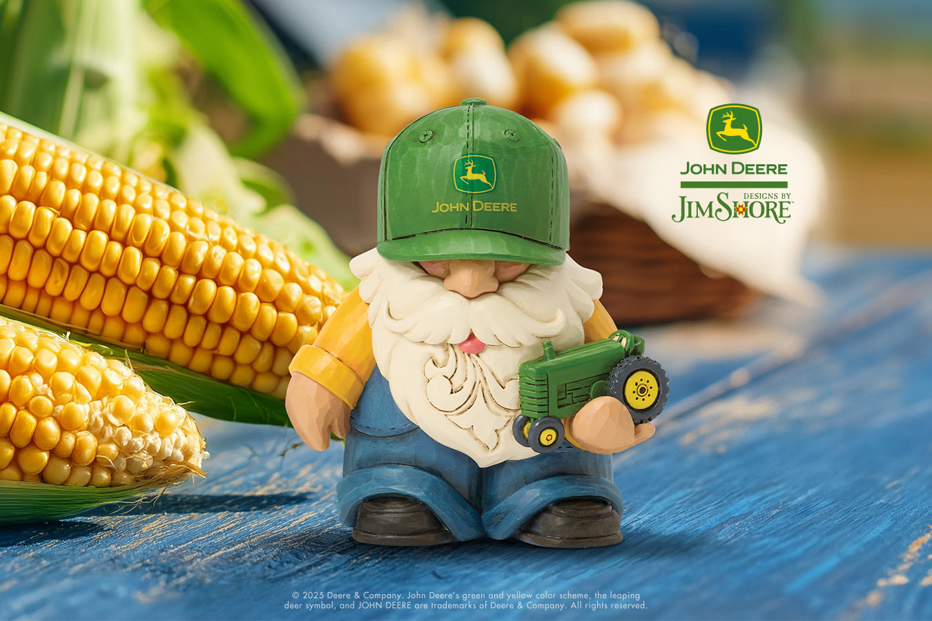Gnome figurine with a John Deere cap and toy tractor next to corn on a blue surface, with Jim Shore branding.