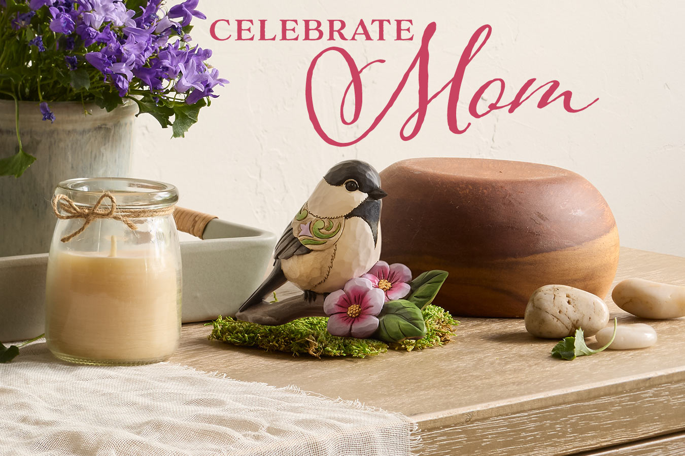 Decorative setup with a candle, bird figurine, and flowers on a wooden surface, celebrating 'Mom'.