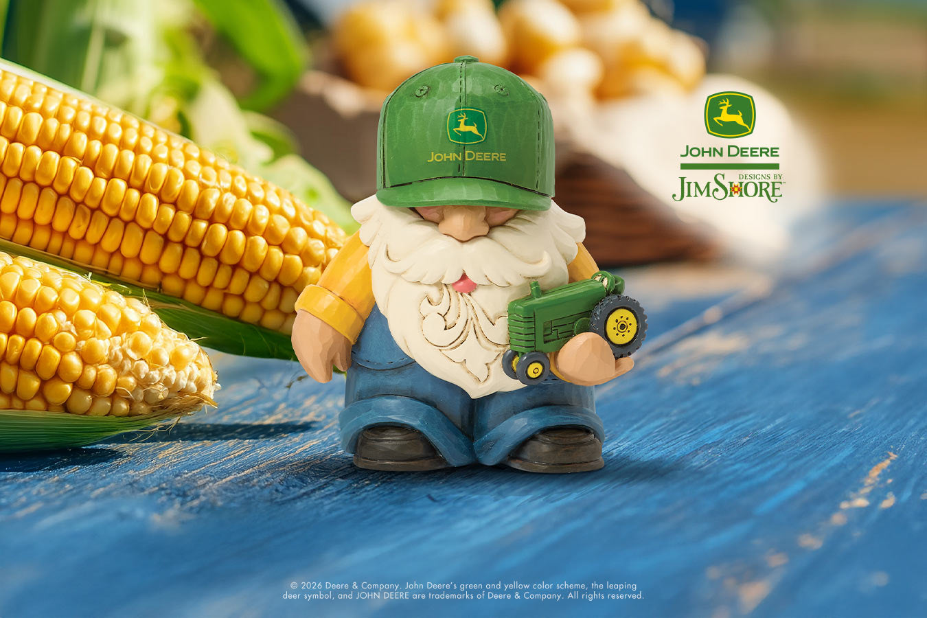Gnome figurine with a green cap and holding a toy tractor next to corn on a blue tarp, with John Deere logos in the background.