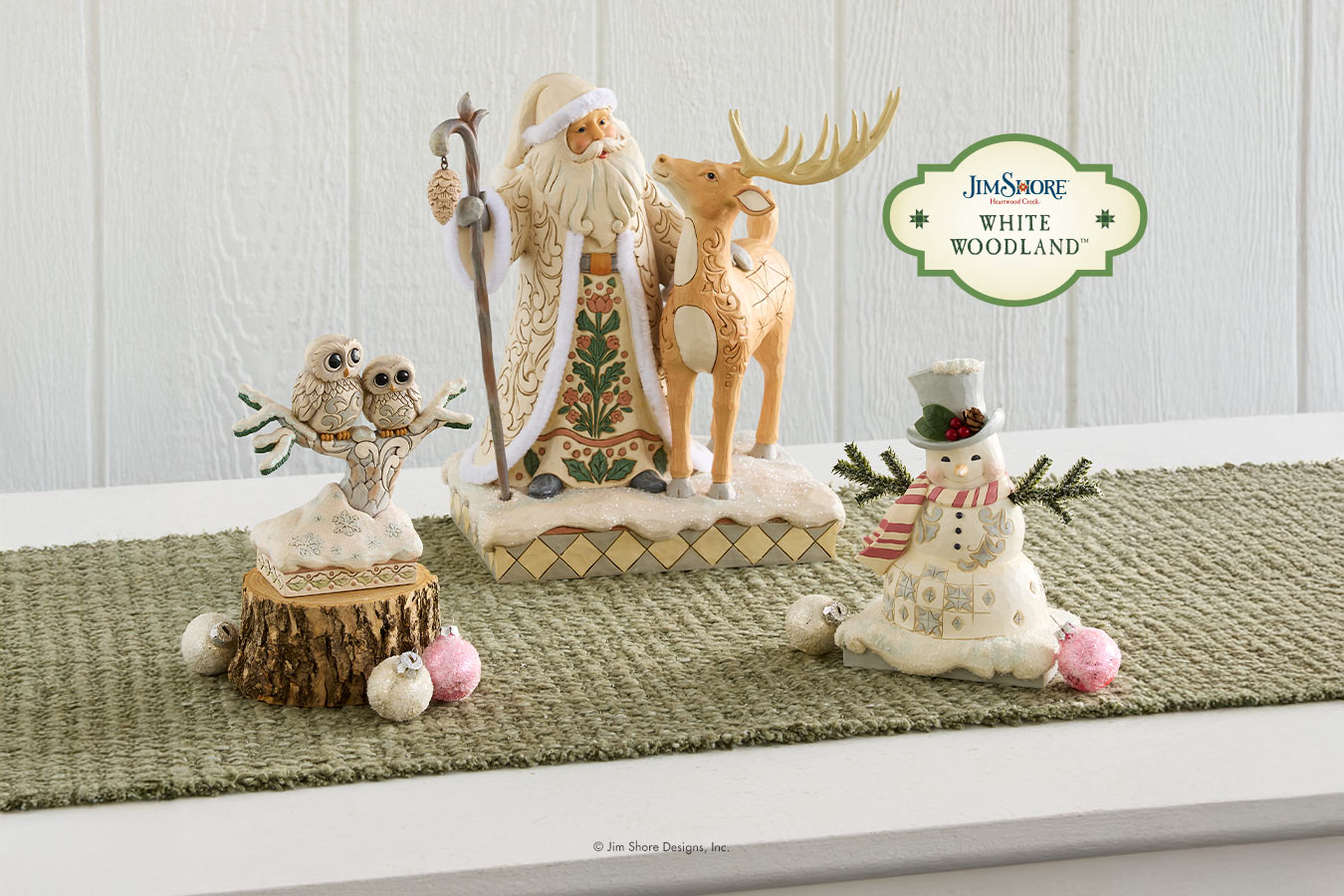 A collection of Jim Stone Designs 'White Woodland' holiday figurines displayed on a green woven runner against a white paneled wall. The set includes a Santa Claus and reindeer figurine, an owl on a tree stump, and a snowman.