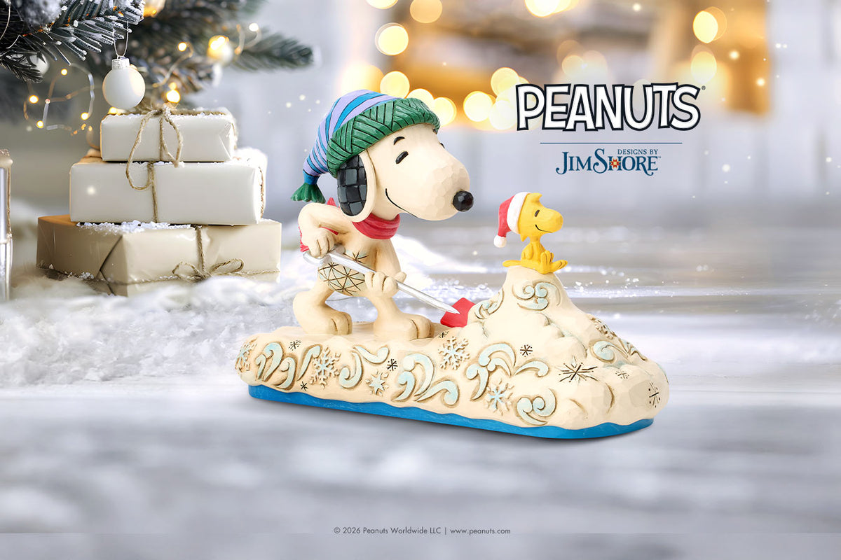 A Peanuts Jim Shore collectible figurine of Snoopy and Woodstock ice skating on a decorated sled, set against a festive blurred holiday background with wrapped gifts and warm bokeh lights. The 'PEANUTS Jim Shore' logo is visible in the upper right.
