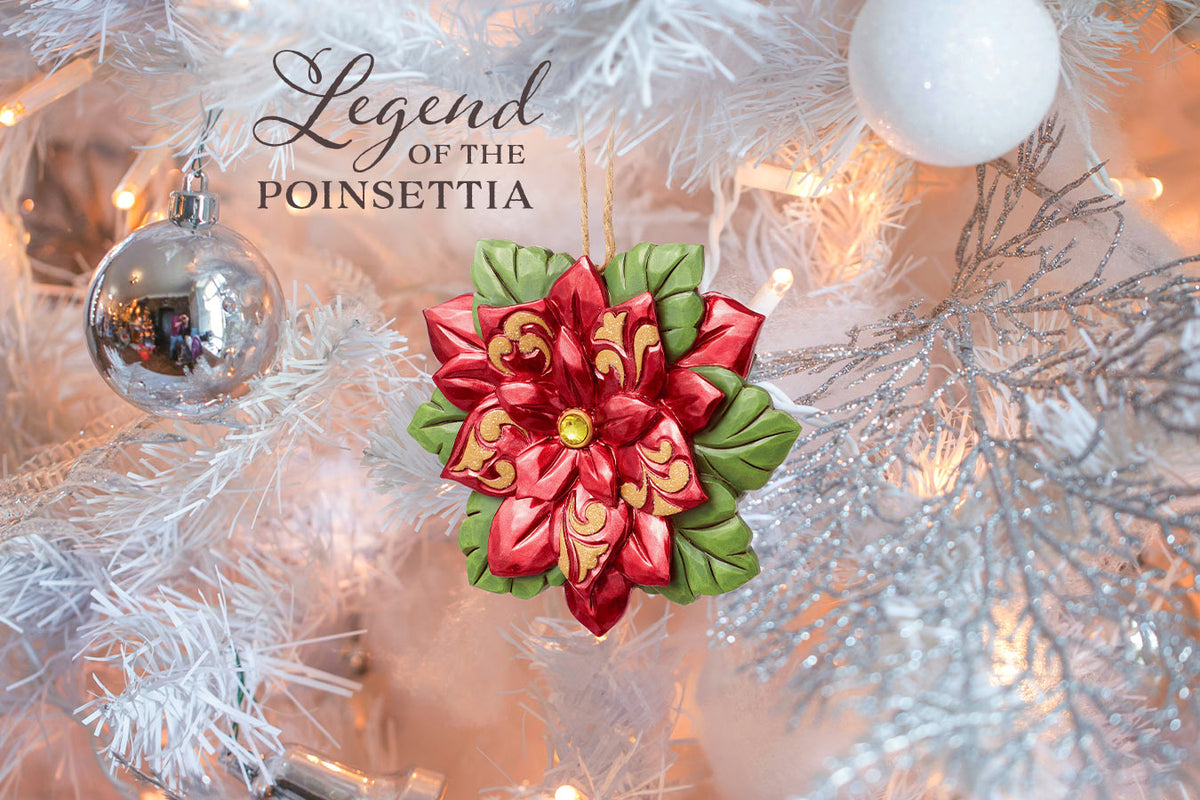 A decorative Christmas ornament shaped like a poinsettia flower hangs on a white flocked Christmas tree. The ornament has red petals with gold detailing, green leaves, and a small gold center.