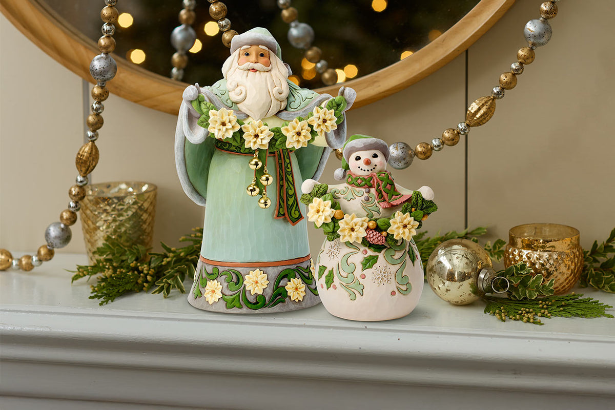 A festive ceramic Christmas ornament set featuring a tall Santa figure in a light green robe holding a wreath and a smaller snowman figurine with floral details, displayed on a mantel with holiday decorations.