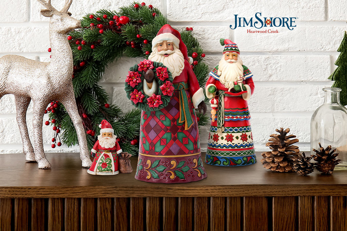 A collection of Jim Shore Christmas figurines displayed on a wooden mantel against a white brick wall. The set includes a large Santa Claus figure in a diamond-patterned red coat holding a red wreath, a smaller Santa figure, a silver glittery reindeer, and a green pine wreath with red berries.