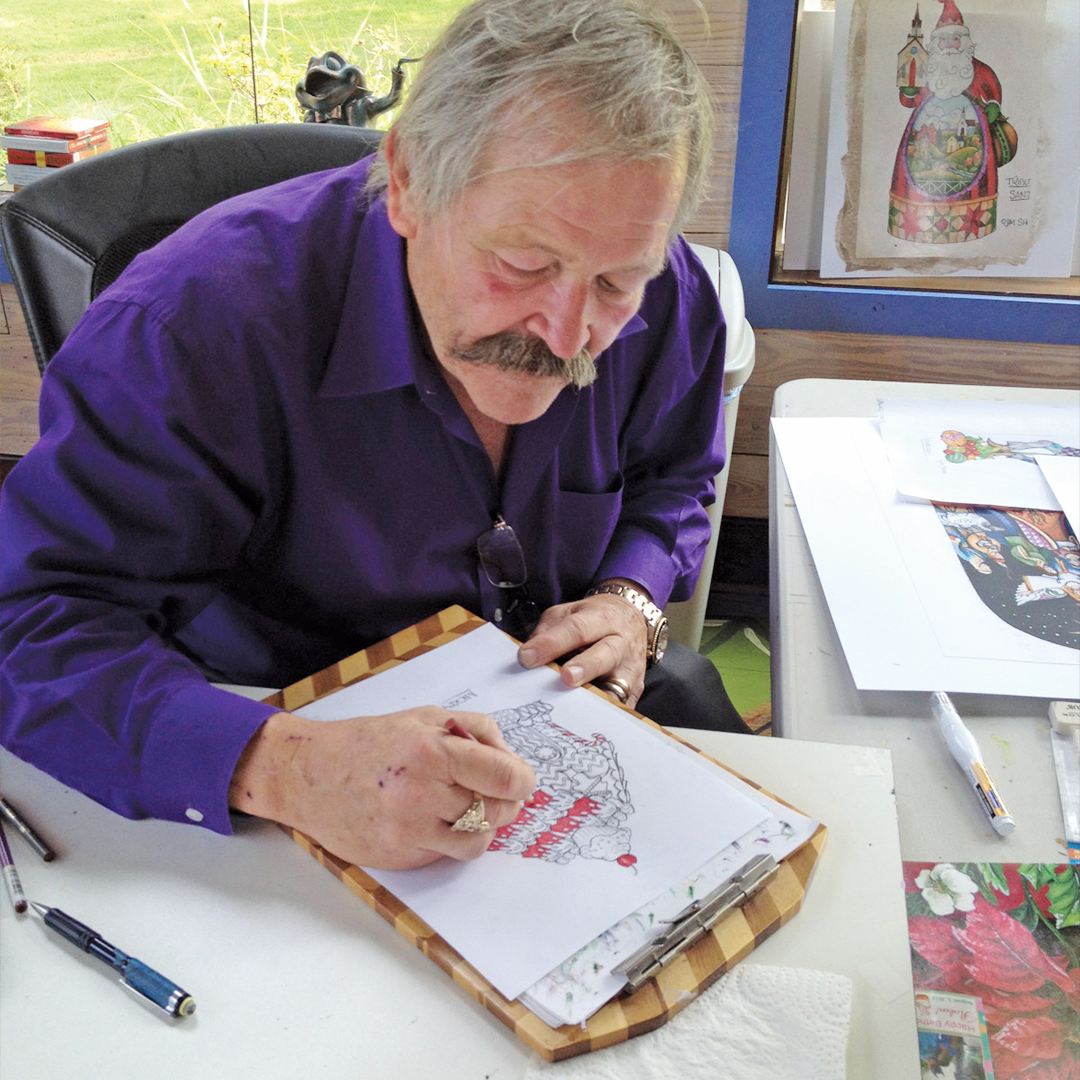 Jim in a purple shirt drawing on paper at a desk with various items around