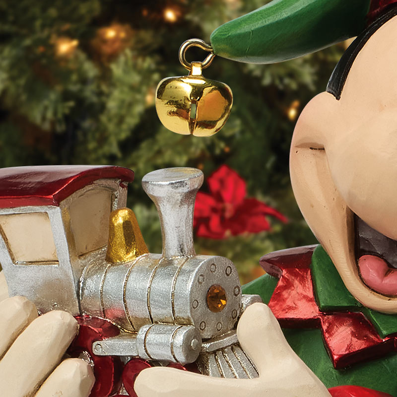 A close-up of a Christmas ornament featuring a silver and red train being held by a stylized character with a green hat and red scarf, with a gold bell hanging above.