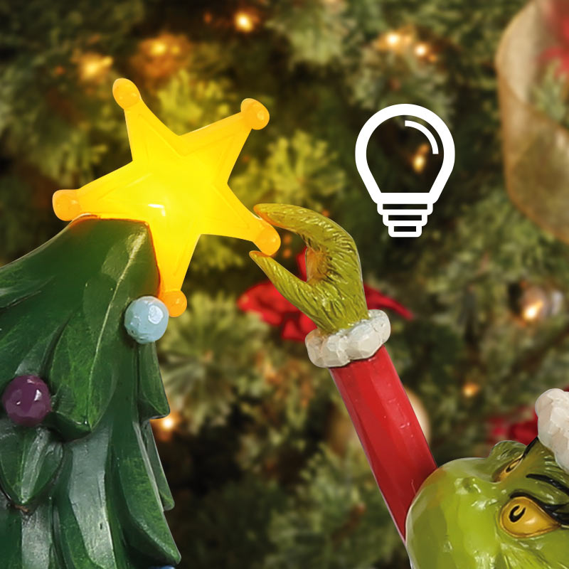 Grinch-themed Christmas ornament close-up showing a green Grinch hand reaching toward a yellow star on a decorated Christmas tree, with a white outline lightbulb icon overlaid on a blurred holiday tree background.
