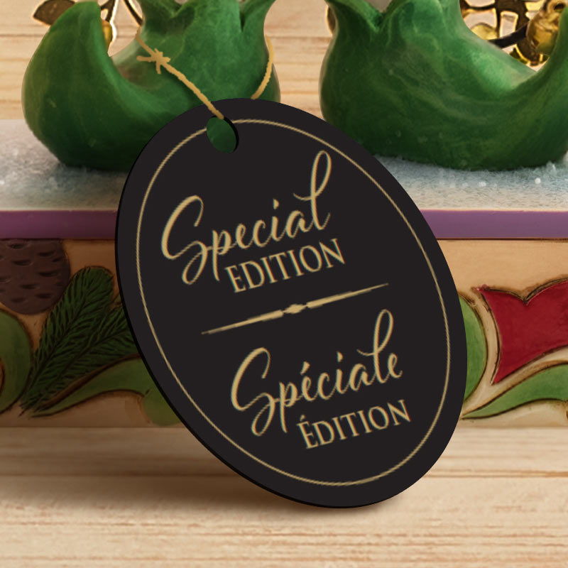 A black oval hanging tag with gold lettering reading 'Special Edition' and 'Spéciale Édition', shown in front of a decorative holiday-themed background with green ornaments and a wooden surface.