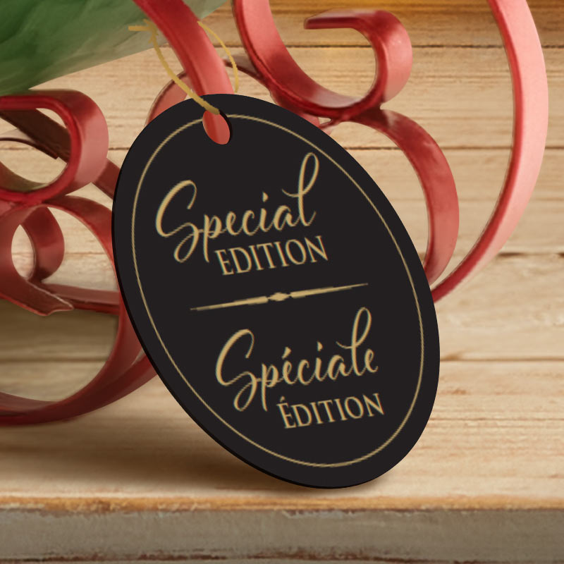 An oval black hanging tag with elegant gold lettering reading 'Special EDITION' and 'Spiciate Édition', photographed on a rustic wooden surface with red ribbon decor in the background.
