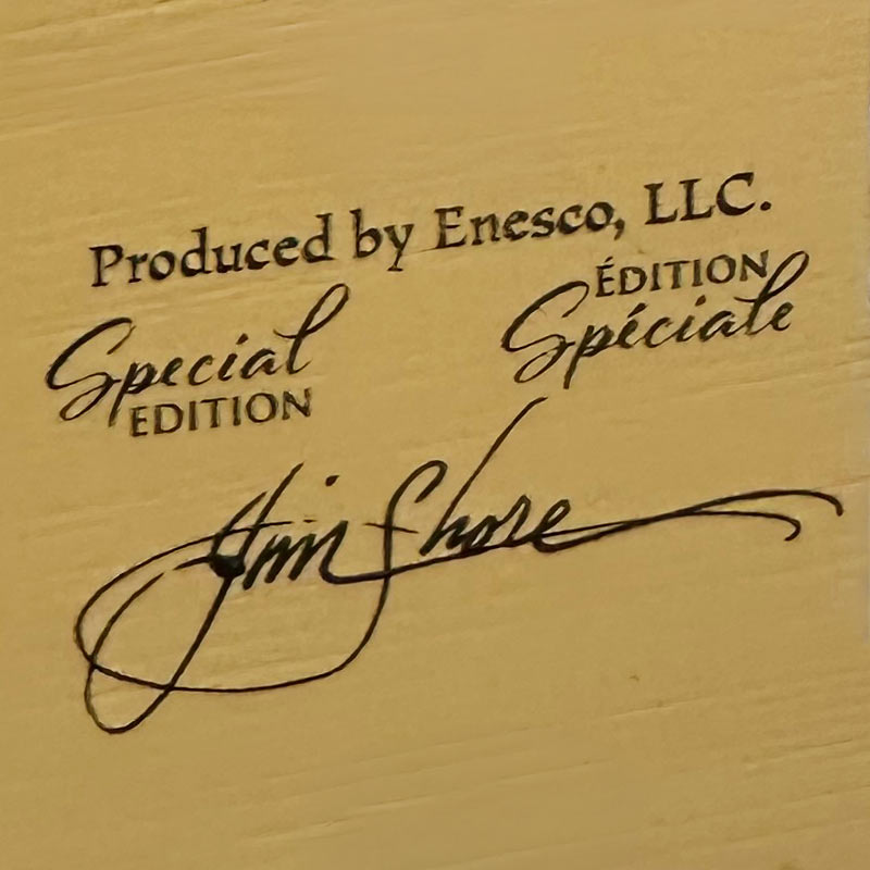 Close-up of a beige or cream textured surface with printed black text reading 'Produced by Enesco, LLC.' and 'Special Edition' / 'Édition Spéciale,' plus a stylized signature that appears to say Jim Shore.