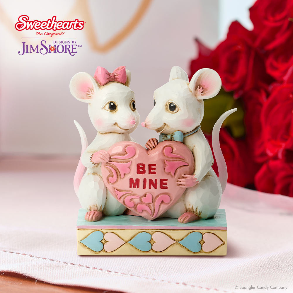 Two mouse figurines holding a heart with 'Be Mine' text, designed by Jim Shore, on a pink background.